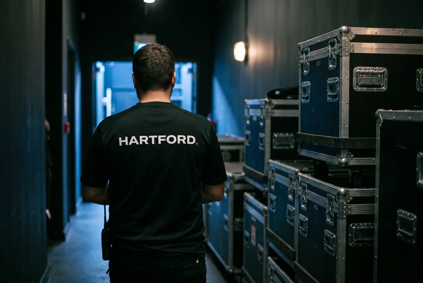 A crew member seen from behind in a black work shirt carrying the Hartford wordmark, walking a backstage corridor lined with road cases.